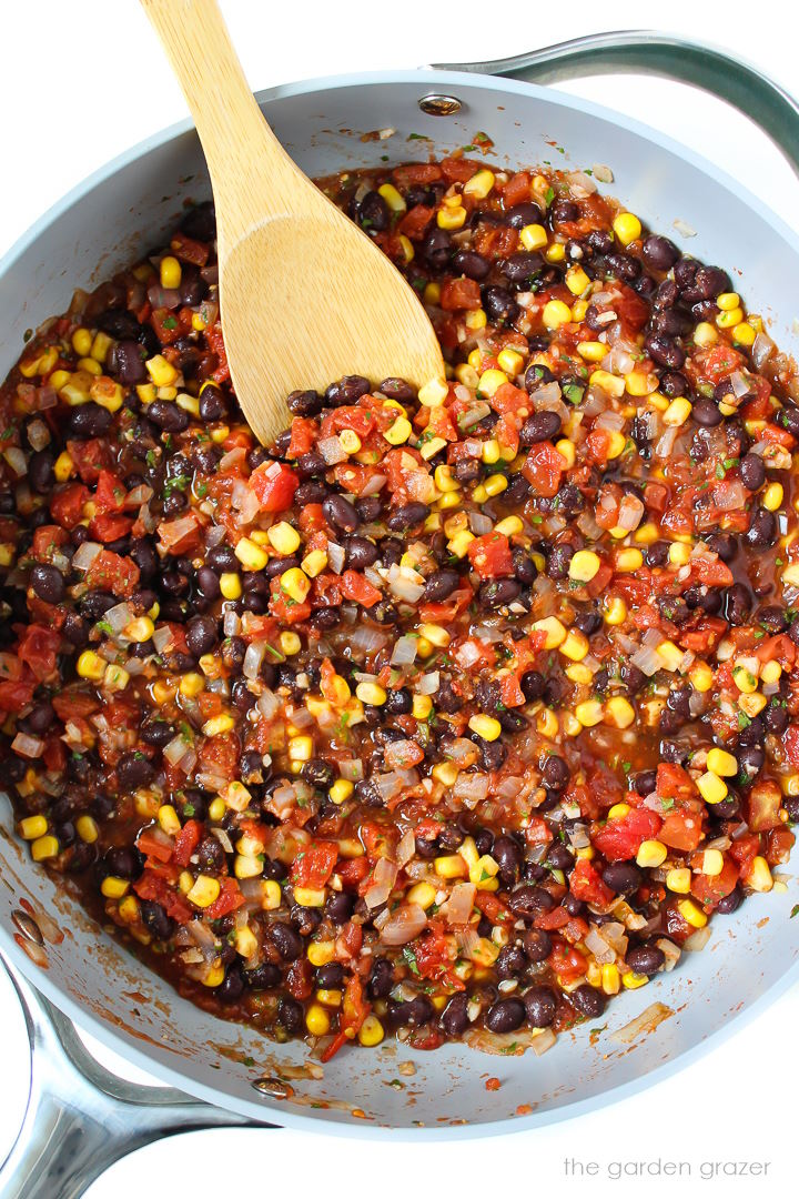 Overhead view of a black bean mixture with tomatoes and corn cooking in a large skillet with wooden spoon