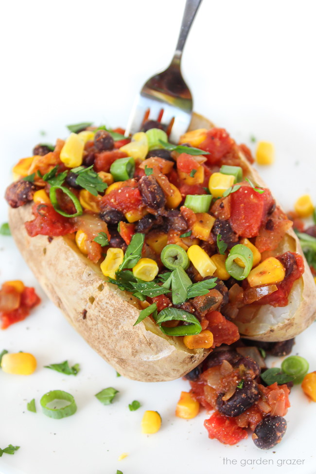 Baked potato topped with black beans, corn, and tomato on a white plate with fork