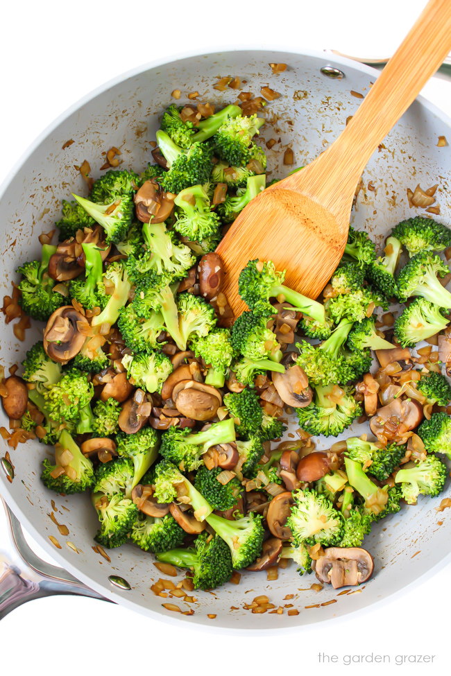 Broccoli and mushrooms cooking in a skillet with wooden spatula