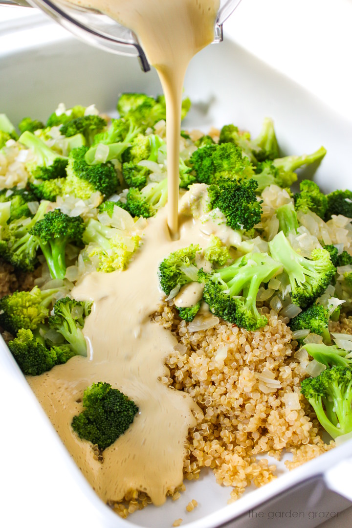 A creamy, cheesy cashew sauce being poured over broccoli quinoa casserole ingredients in a large white baking dish