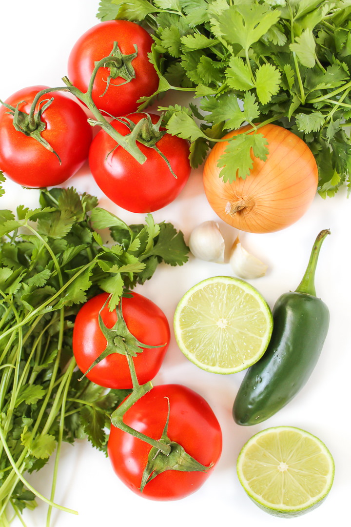 Fresh cilantro, tomato, onion, garlic, lime, and jalapeno on a white table for pico de gallo