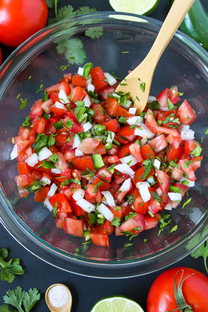 Overhead view of preparing salsa fresca in a large glass bowl with wooden spoon