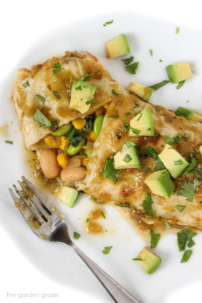 Vegan enchiladas verde on a white plate with avocado and fork