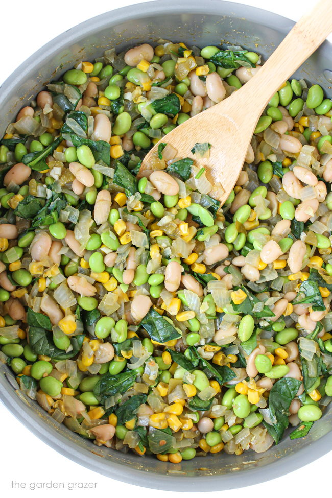 Onion, spinach, soybeans, and sweet corn cooking in a skillet with wooden spoon