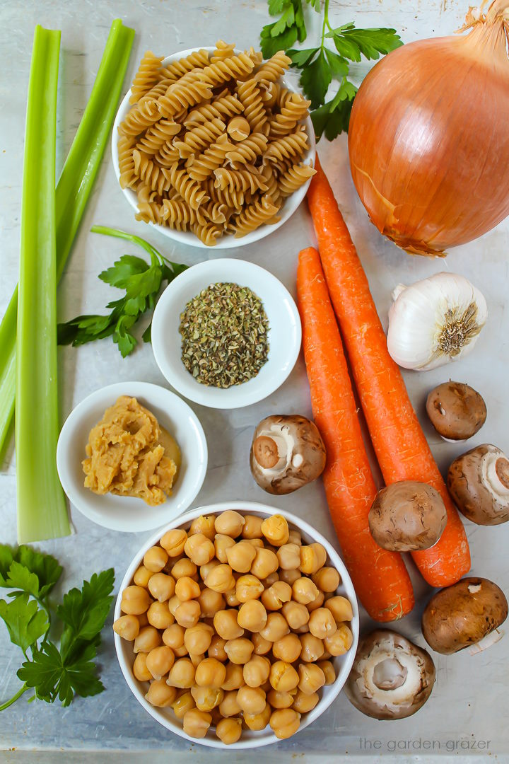 Fresh carrot, mushrooms, celery, pasta, garbanzo beans, onion, and herb ingredients laid out on a metal tray