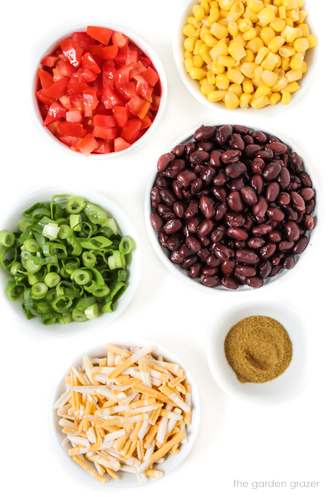 Beans, sweet corn, tomatoes, green onions, cumin, and vegan cheese ingredients on a white table