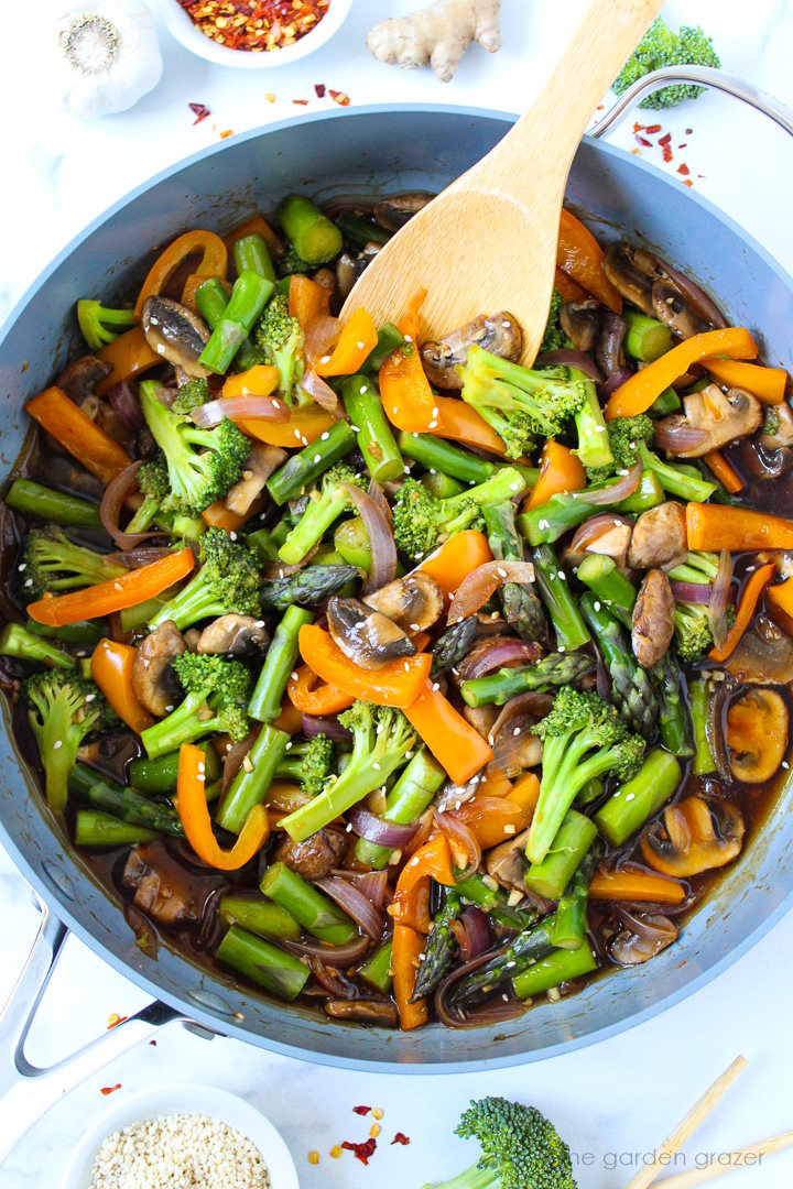 Overhead view of saucy stir fry vegetables in a large skillet with wooden stirring spoon