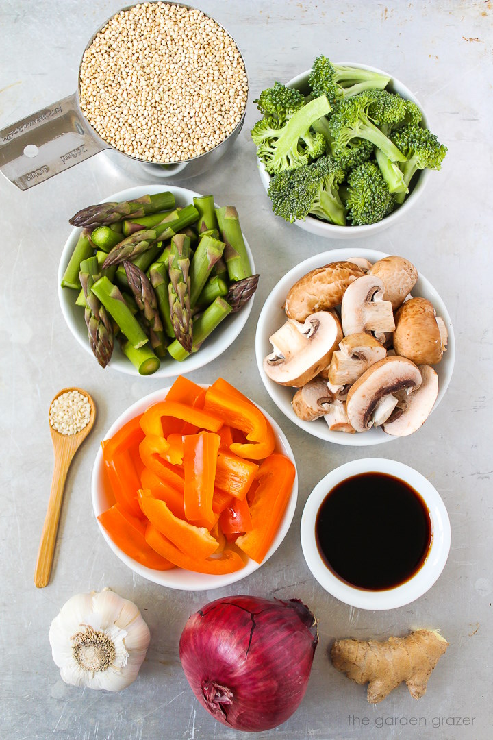 Overhead view of fresh broccoli, asparagus, mushrooms, bell pepper, red onion, tamari, and ginger ingredients laid out on a metal tray
