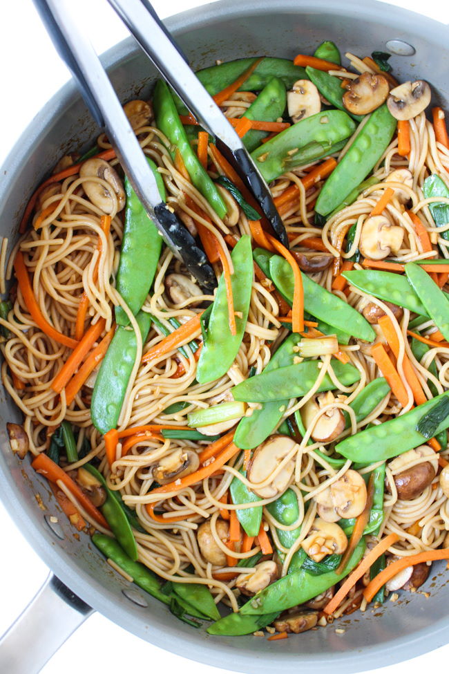 Noodles and vegetables cooking in a large skillet with tongs