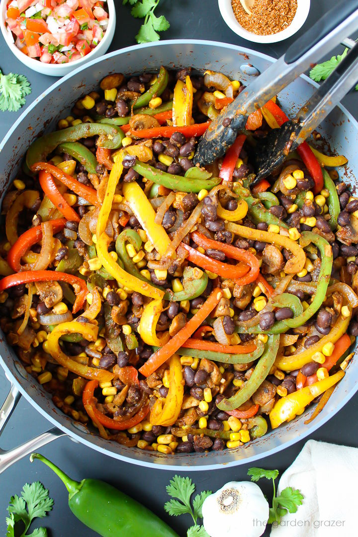 Overhead view of corn and black bean fajitas cooking in a large pan with tongs for stirring