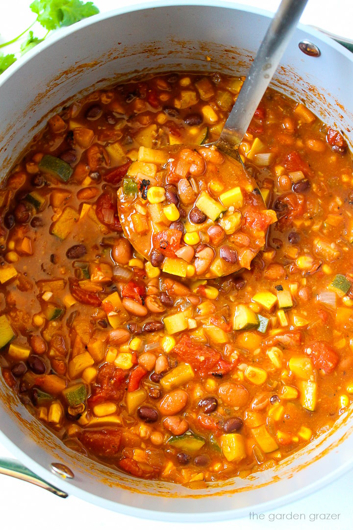 A soup ladle lifting up summer vegetable chili from a large pot as it cooks