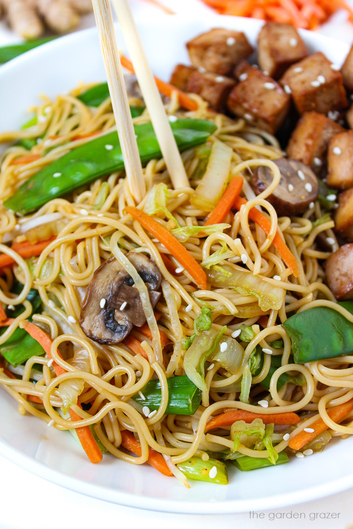 Close up view of vegan vegetable lo mein served on a white plate with tofu and chopsticks