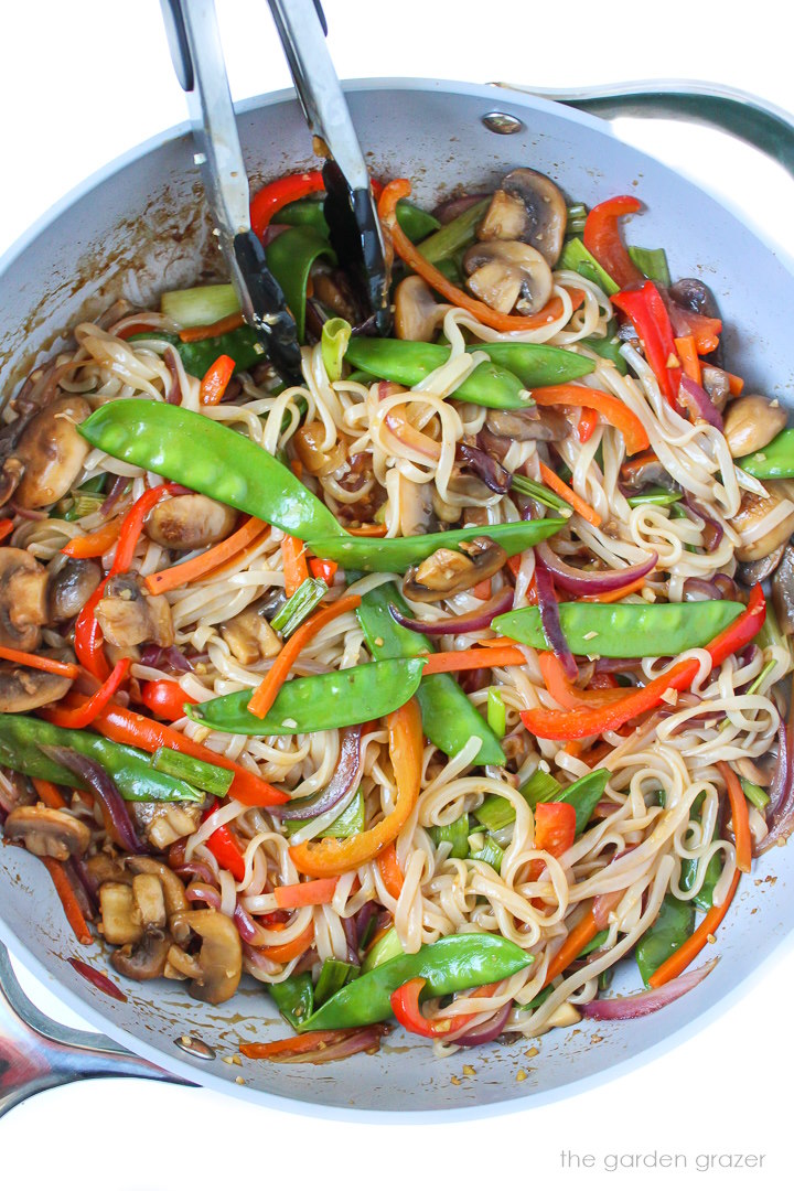Vegan vegetable lo mein cooking in a large skillet with tongs