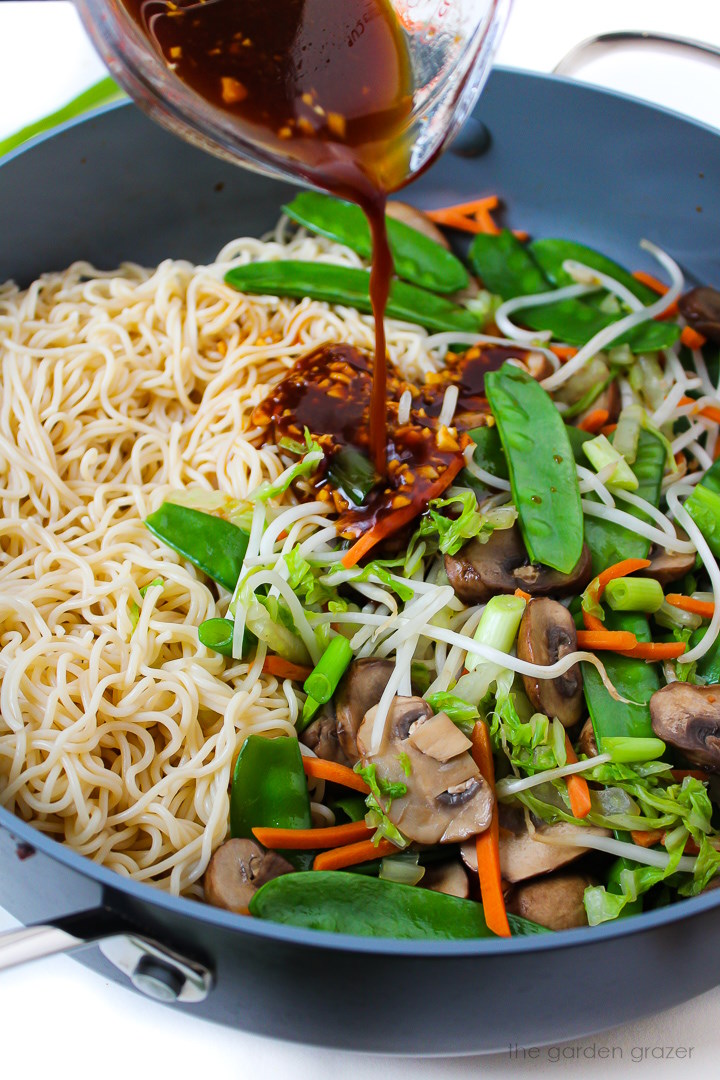 A savory sauce being poured over noodles and veggies in a large cooking skillet