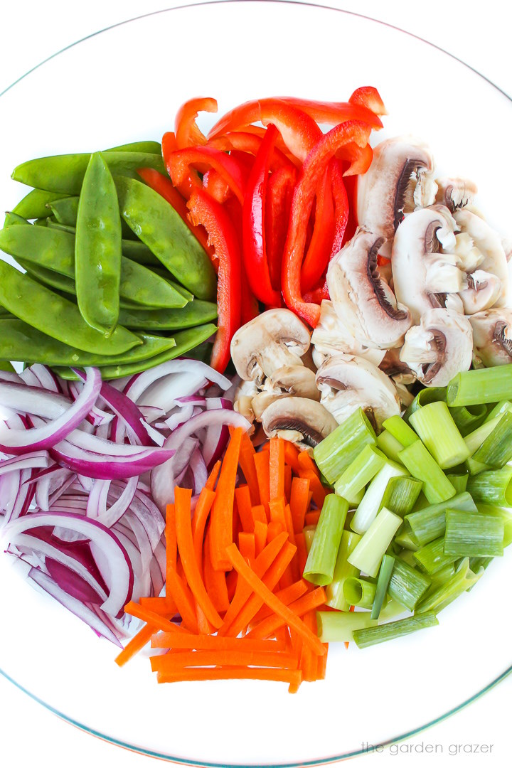 Chopped raw veggies like mushrooms, bell pepper, onion, peas, and carrots in a large glass bowl