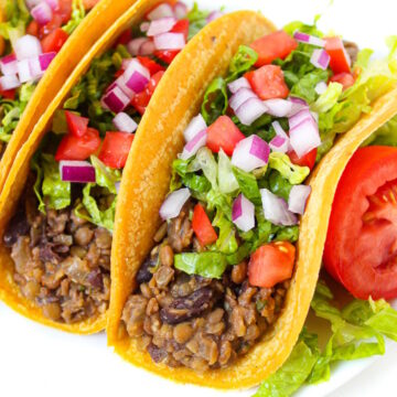 Three black bean lentil tacos on a white plate topped with lettuce, tomato, and onion