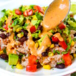 A spoon drizzling creamy chipotle sauce over a vegan black bean burrito bowl on a white plate