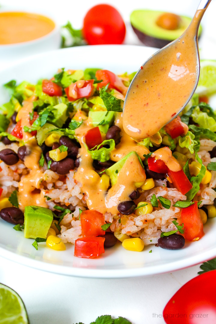 A spoon drizzling creamy chipotle sauce over a vegan black bean burrito bowl on a white plate