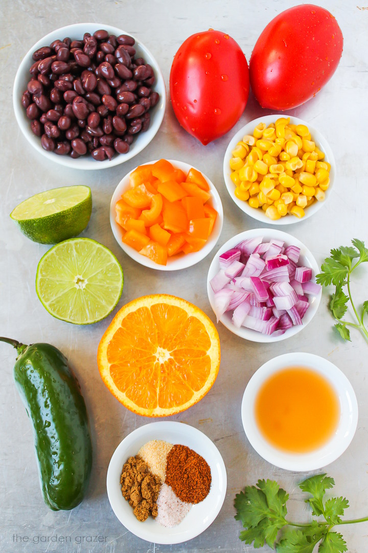 Roma tomatoes, sweet corn, red onion, bell pepper, fresh orange, lime and spice ingredients laid out on a metal tray