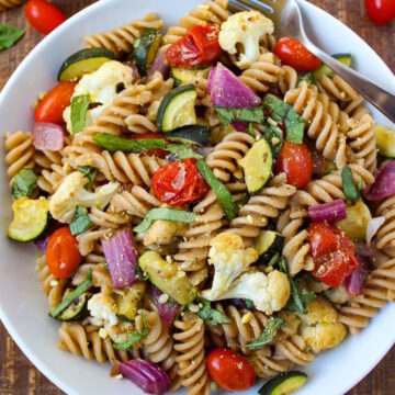 A white bowl of roasted vegetable pasta on a wooden table with serving fork