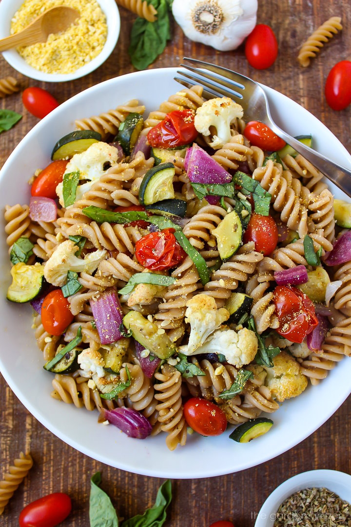 A white bowl of roasted vegetable pasta on a wooden table with serving fork