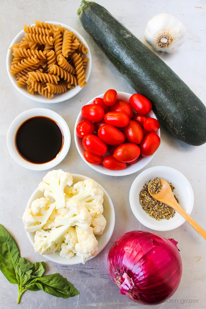Zucchini, grape tomatoes, garlic, cauliflower, red onion, fresh basil, and seasoning ingredients laid out on a metal tray