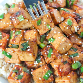 Fork piercing a piece of Asian Style Garlic Tofu from bowl with rice