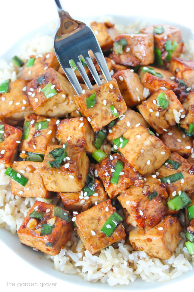 Fork piercing a piece of Asian Style Garlic Tofu from bowl with rice