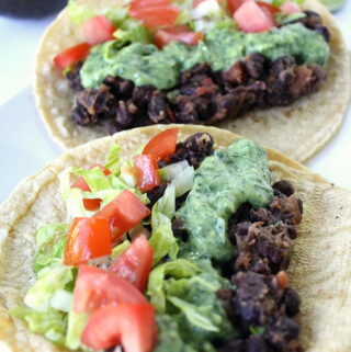 Two black bean tacos on a plate topped with cilantro sauce and tomatoes