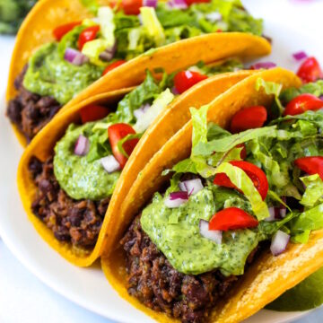Black bean tacos served on a white plate topped with creamy avocado cilantro-lime sauce