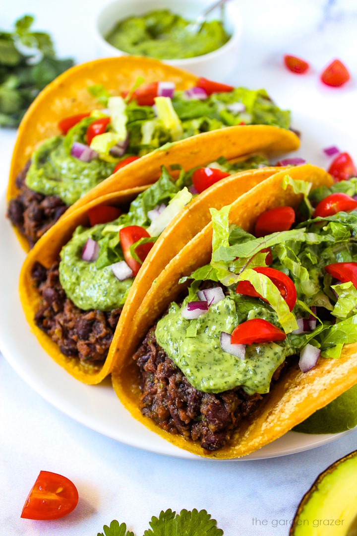 Black bean tacos served on a white plate topped with creamy avocado cilantro-lime sauce