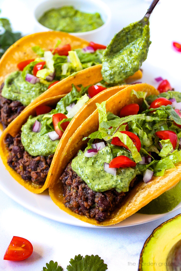 A spoon drizzling avocado cilantro sauce over prepared black bean tacos on a white plate