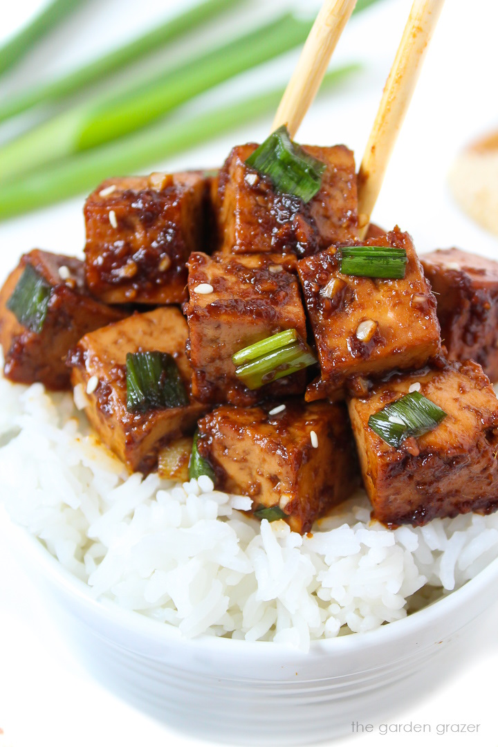 Chopsticks picking up a cube of Asian Garlic Tofu from a bowl of rice