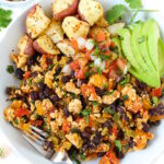 Overhead view of southwest tofu scramble in a white bowl with roasted potatoes, salsa, and avocado slices