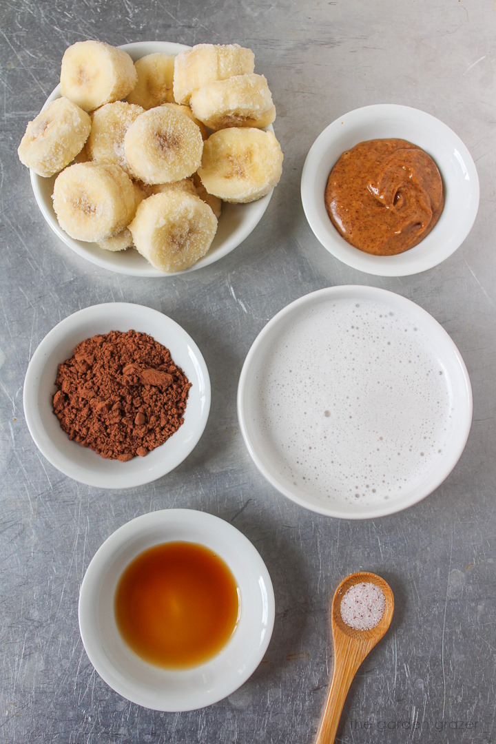 Frozen banana coins, dairy-free milk, cocoa powder, almond butter, and vanilla ingredients laid out on a metal tray