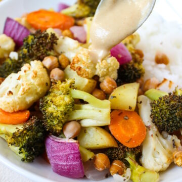 A spoon drizzle tahini sauce over roasted vegetables and chickpeas in a white bowl