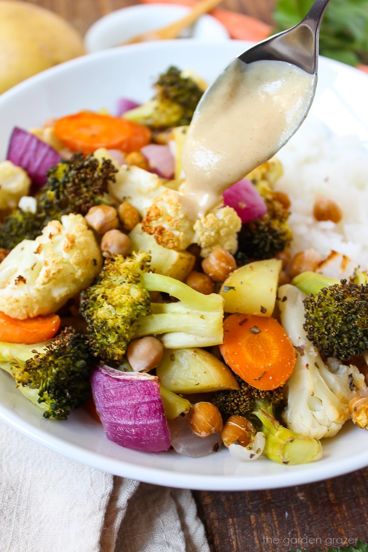 A spoon drizzle tahini sauce over roasted vegetables and chickpeas in a white bowl
