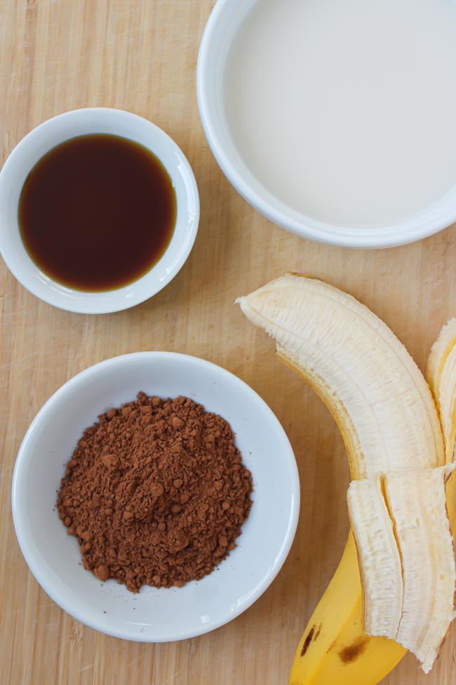 Dairy-free milk, banana, cacao powder, and maple syrup ingredients on a bamboo board