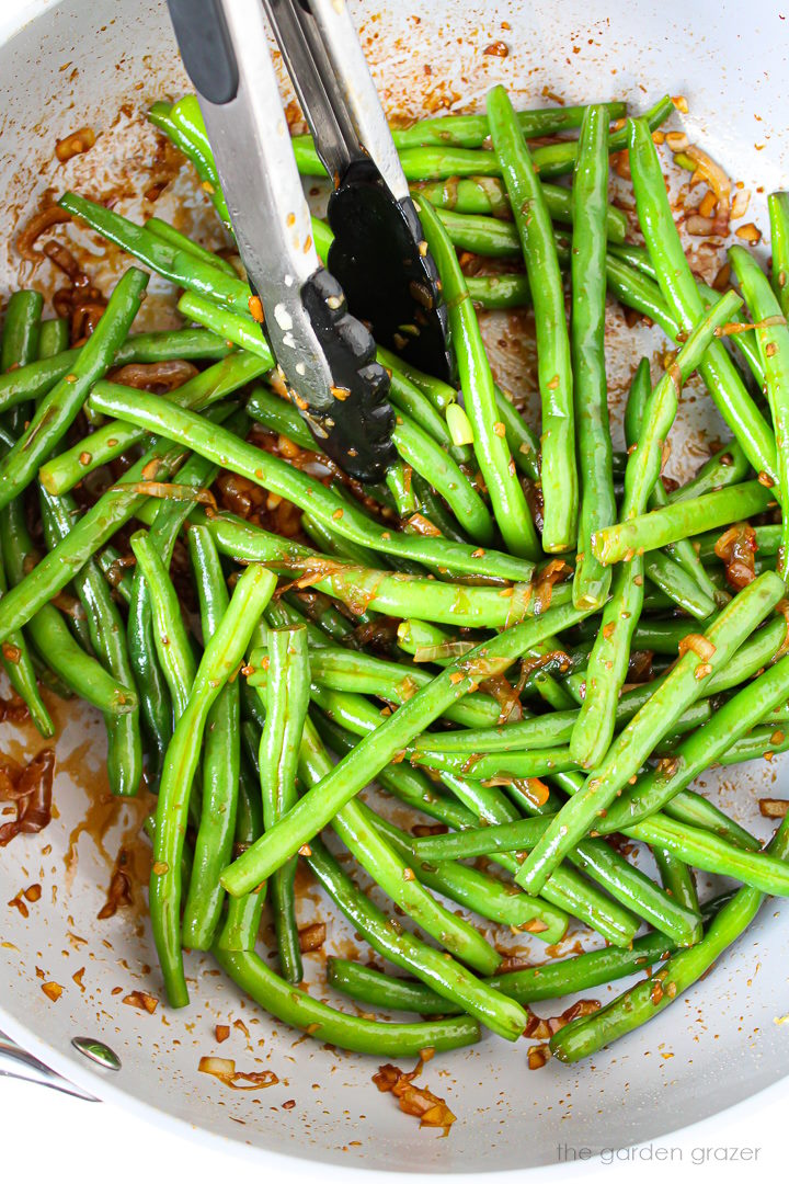 Overhead view of Asian garlic green beans cooking in a large pan with tongs for stirring