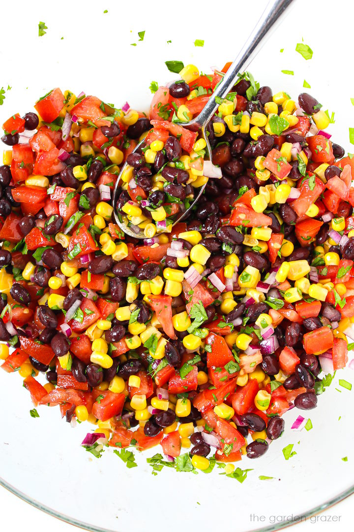 Mixing together black bean and corn salsa with a metal spoon in a large glass bowl