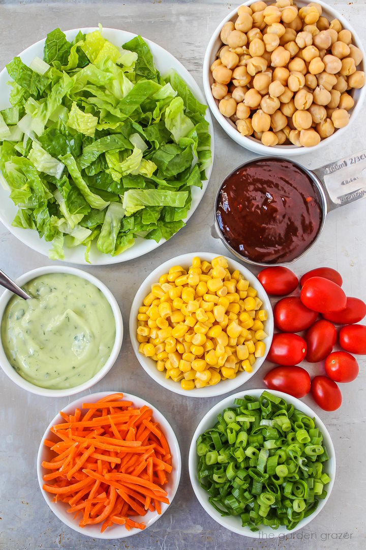 Chopped romaine lettuce, garbanzo beans, sweet corn, BBQ sauce, carrots, and tomato ingredients laid out on a metal tray