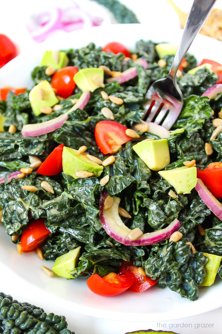 A fork piercing Marinated Kale Salad served on a white plate with avocado and tomato
