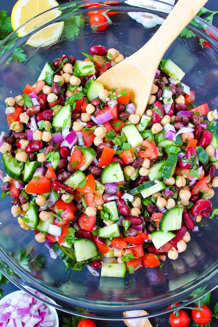 Overhead view of preparing Mediterranean Three Bean Salad in a large bowl with wooden stirring spoon