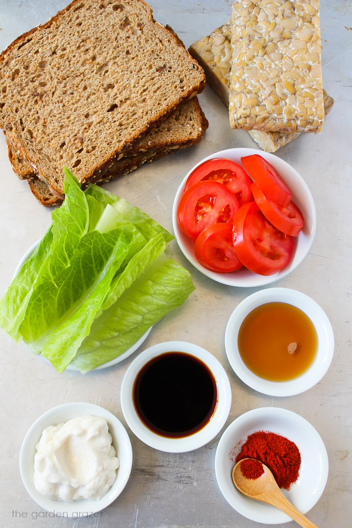 Overhead view of bread slices, tomatoes, romaine lettuce, tamari, smoked paprika, and mayo ingredients laid out on a metal tray
