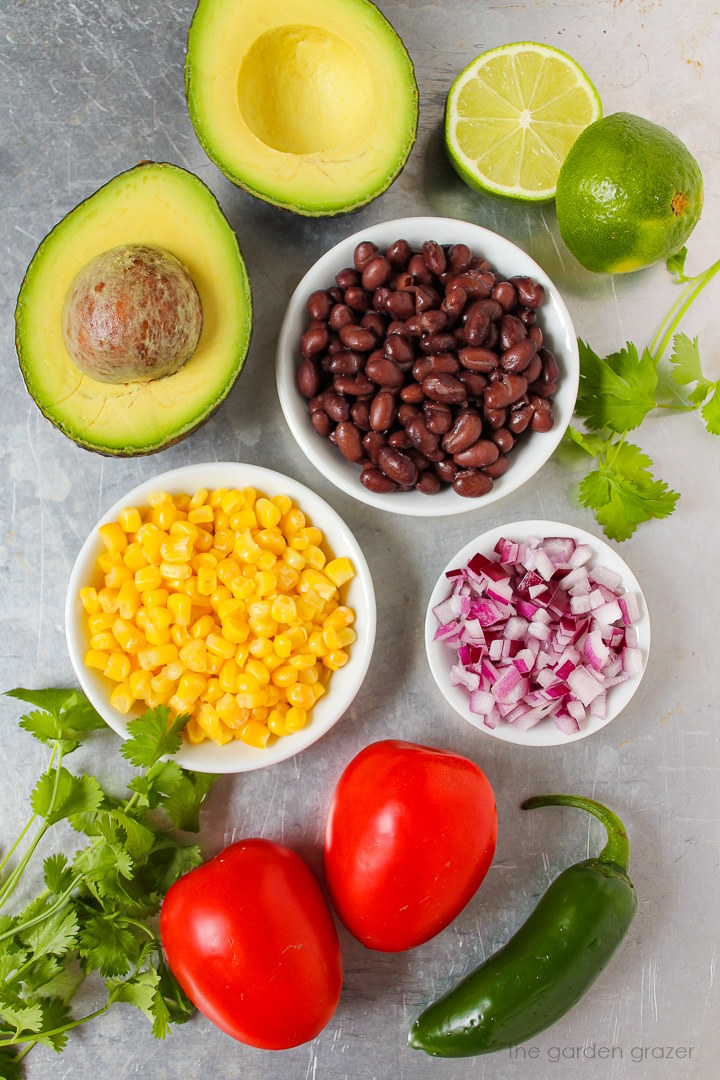 Avocado, black beans, sweet corn, red onion, roma tomato, and fresh lime ingredients laid out on a metal tray