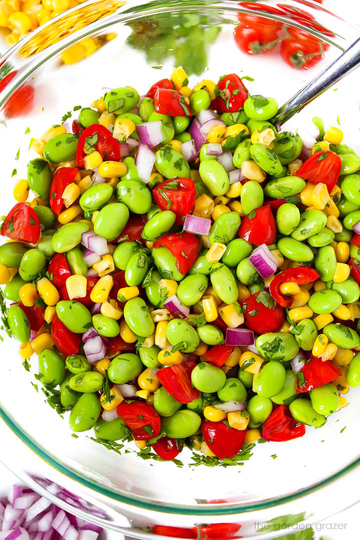 Edamame corn salad with tomato and cilantro mixed together in a large glass bowl with spoon