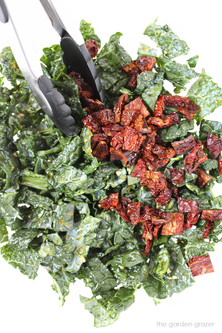Chopped kale and sun-dried tomatoes in a glass bowl with tongs