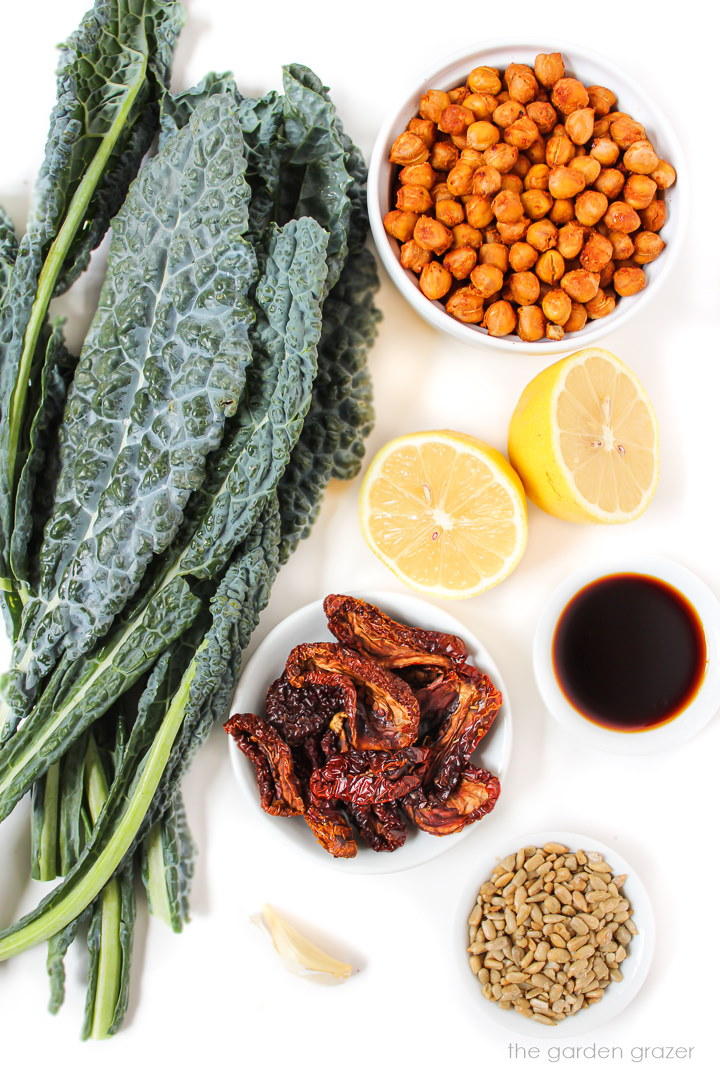 Fresh kale, roasted chickpeas, lemon, and sun-dried tomato ingredients laid out on a white table