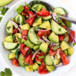 Cucumber tomato avocado salad with lemon and cilantro in a white bowl with serving spoon