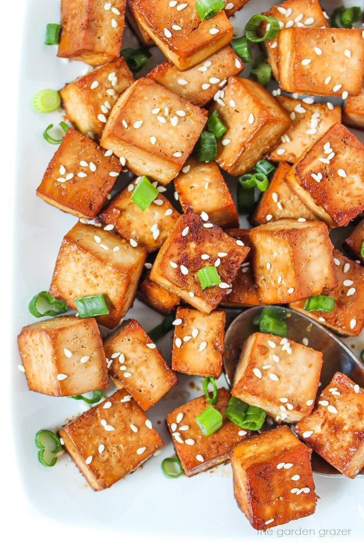 Overhead view of easy baked savory tofu cubes on a white plate with sesame seeds and green onions